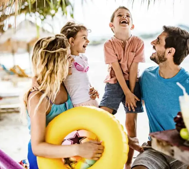 Lachende Familie am Strand mit gelbem Schwimmring und Getränken.