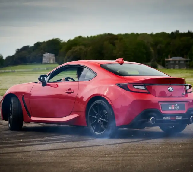 Czerwona Toyota 86 GR w poślizgu, z opon dymiących na asfalcie. W tle zielone pola i drzewa.