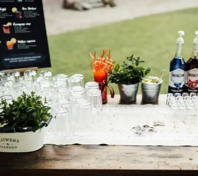 Stół z kieliszkami, butelkami syropów i miętą, gotowy na przygotowanie drinków na wesele. Przepisy na drinki są widoczne na tablicy w tle.