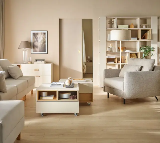 Jasny salon z sofą, fotelem i stolikiem kawowym na kółkach. Na półkach regału stoją dekoracje, książki i lampa. Wersaliki dodają elegancji.