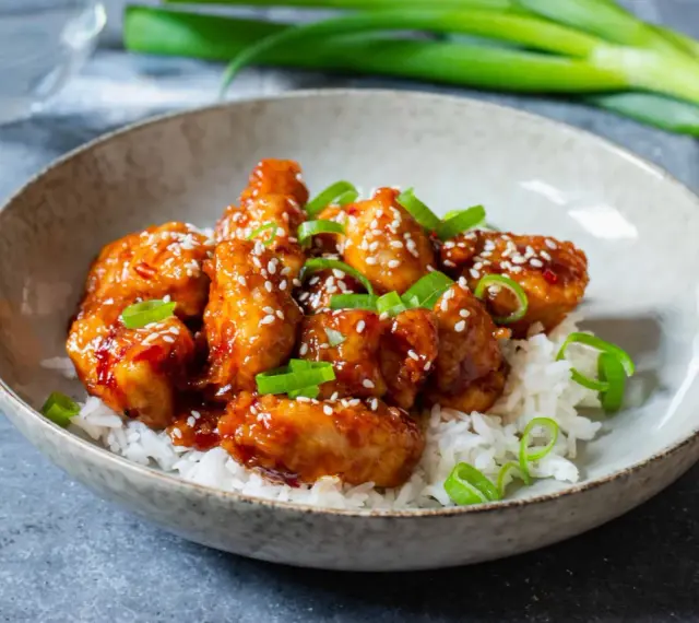 Krispig koreansk kyckling med sesamfrön och salladslök serveras på en bädd av vitt ris i en skål.