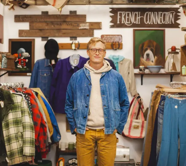 Mężczyzna w sklepie z odzieżą vintage, gdzie wisi szyld "FRENCH CONNECTION". W tle wiszą ubrania i dekoracje.