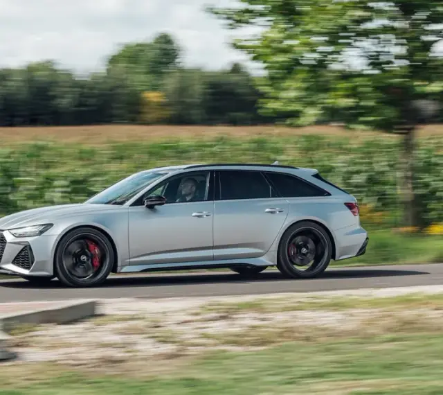 Srebrne Audi RS6 Avant pędzi po wiejskiej drodze, otoczone zielenią pól i drzew.