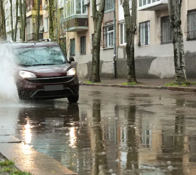Ciemny samochód wpada w kałużę, powodując potężny **aquaplaning**. Woda rozpryskuje się na boki, a odbicia budynków i drzew tańczą na mokrej nawierzchni.