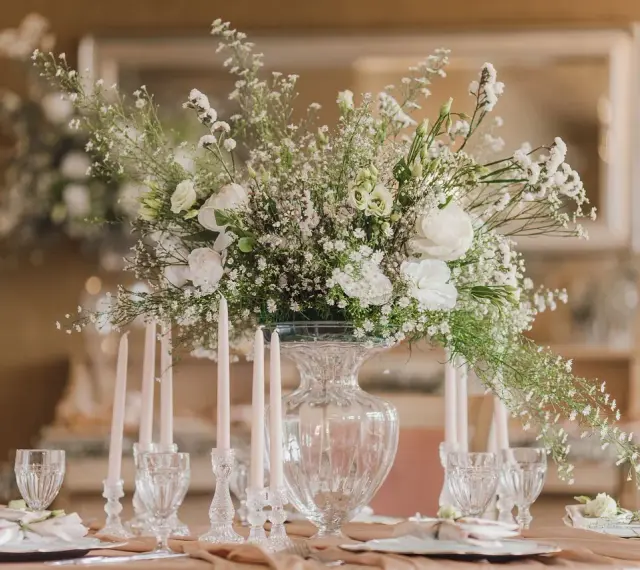 Eleganckie ustawienie stołów na 40 osób. Centralny punkt stanowi bukiet białych kwiatów w szklanym wazonie, otoczony świecami i kryształowymi kieliszkami.