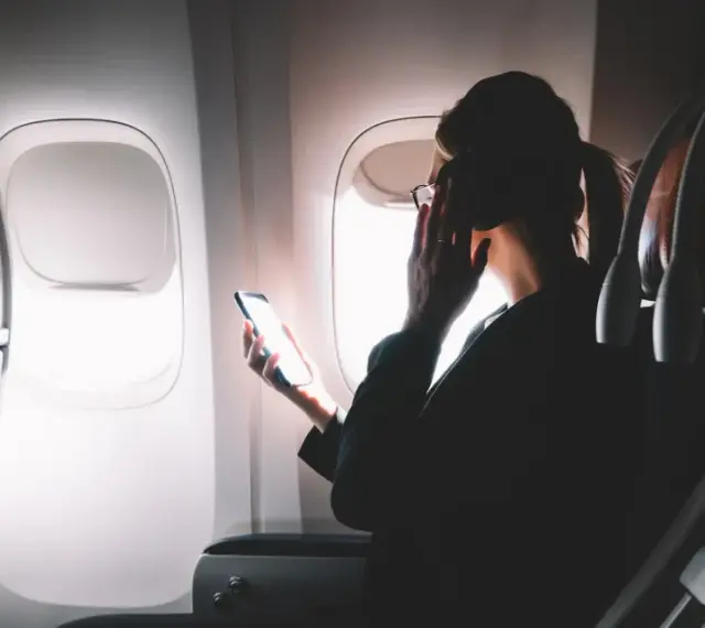 Kobieta w okularach trzyma telefon, zastanawiając się, czy w samolocie jest wifi. Obok ekran rozrywki.