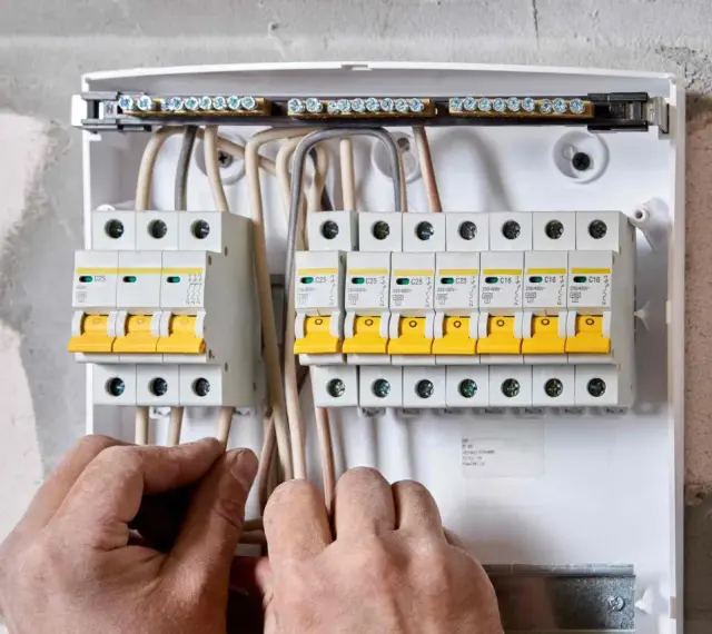 Ręce elektryka podłączają przewody do skrzynki z bezpiecznikami. Widać wlz, czyli rozdzielnicę z wyłącznikami nadprądowymi.
