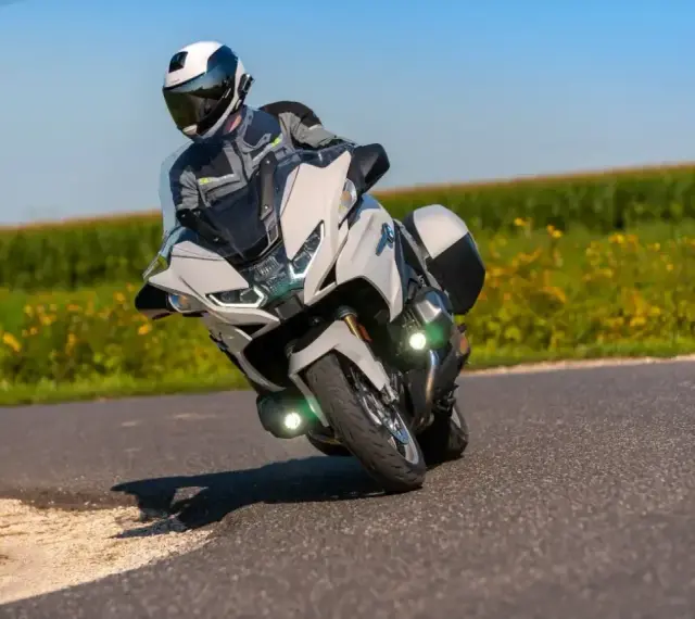 Motocyklista w kasku i kombinezonie pochyla się na zakręcie na białym motocyklu BMW R 1250 RT, przemierzając malowniczą drogę wśród pól.
