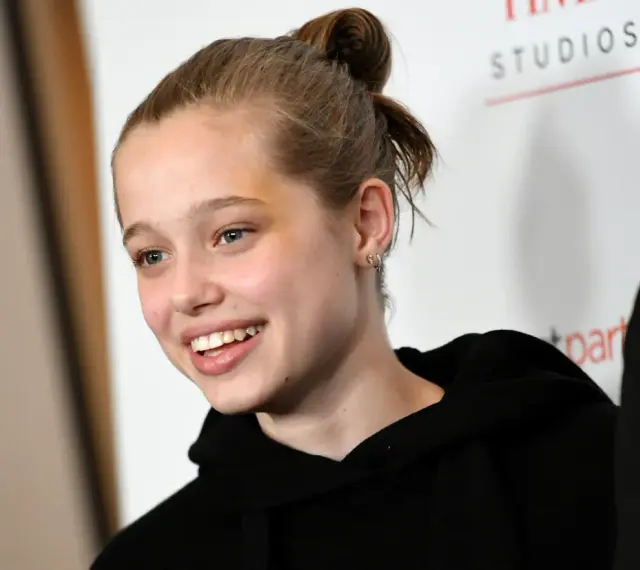 Uśmiechnięta Shiloh Jolie Pitt z włosami upiętymi w kok, w czarnej bluzie z kapturem.