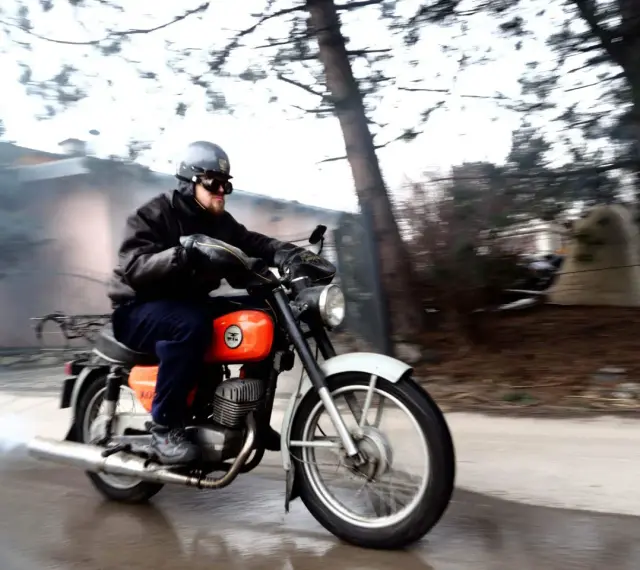 Mężczyzna w kasku i goglach pędzi na pomarańczowym motocyklu WSK 125. Widać dym z rury wydechowej, sugerujący dużą prędkość maksymalną.