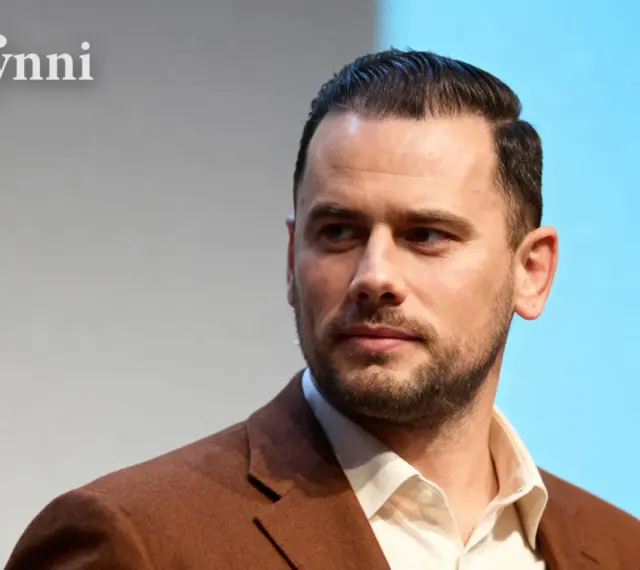 Grzegorz Collins, znany z programów telewizyjnych, w eleganckim brązowym garniturze. Jego wiek jest trudny do określenia, ale prezentuje się dojrzale.