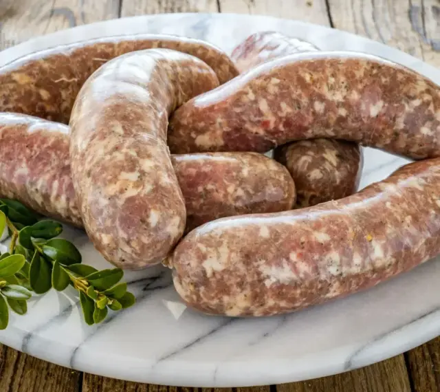 Kilka surowych kiełbasek białych parzonych ułożonych na marmurowej desce, ozdobione gałązką zielonych liści.