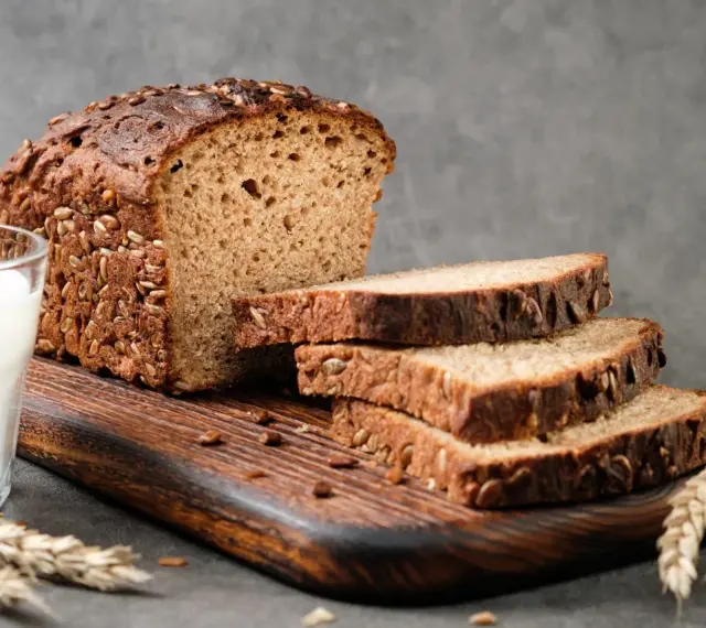 Chleb proteinowy z ziarnami, pokrojony na kromki, obok szklanka mleka i kłosy zboża. Idealny na zdrowe śniadanie.
