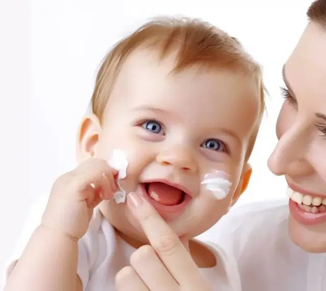 Uśmiechnięty bobas z białym kremem na policzkach, mama delikatnie go dotyka. Czy sudocrem wysusza? Nie w tym przypadku, krem pielęgnuje delikatną skórę.