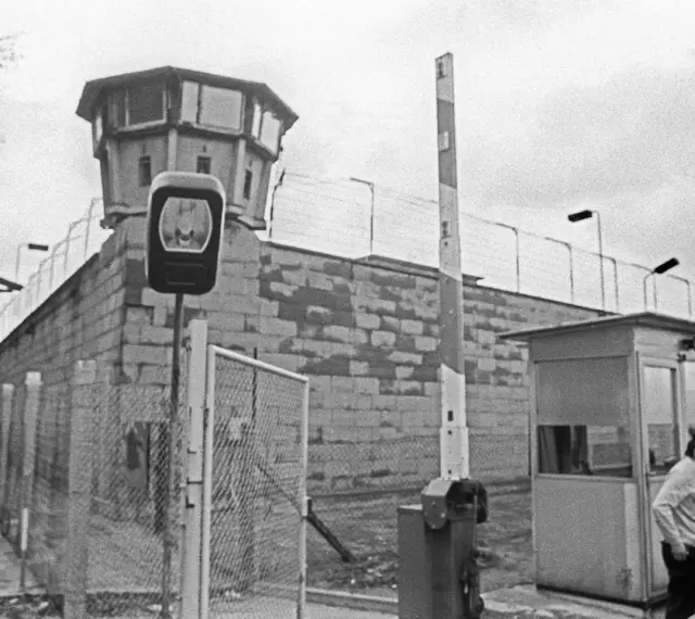 Wachturm und Grenzbefestigung der deutschen demokratischen Republik. Ein Wachmann steht neben einer Schranke und einem Kontrollhäuschen.