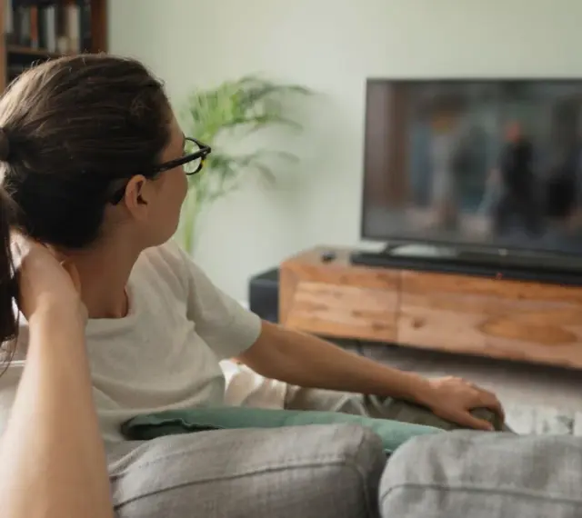 Kobieta w okularach relaksuje się na kanapie, oglądając telewizor. Idealna jaka odległość od telewizora 65 cali?