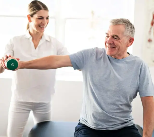 Mann hebt Hantel unter Anleitung einer Therapeutin. Physiotherapie nach Bobath hilft bei der Rehabilitation.