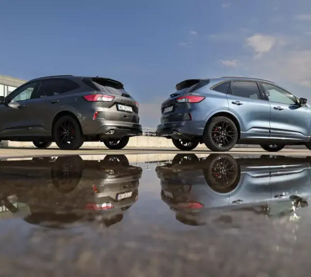 Dwa Fordy Kuga, jeden szary, drugi niebieski, stoją obok siebie na parkingu. Ich odbicia w kałuży tworzą surrealistyczny obraz. Oba modele to wersje hev.