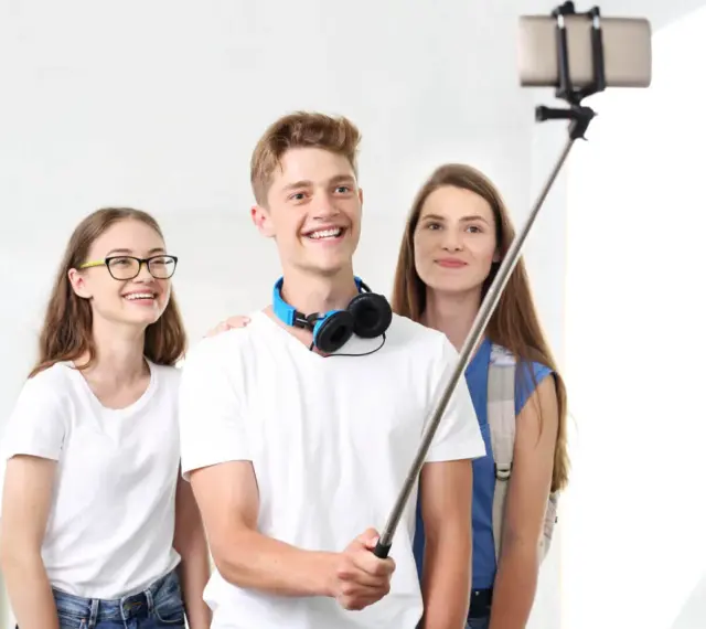 Trzech przyjaciół robi sobie selfie na kijku. Chłopak z przodu uśmiecha się szeroko, pokazując, jak robić zdjęcia.