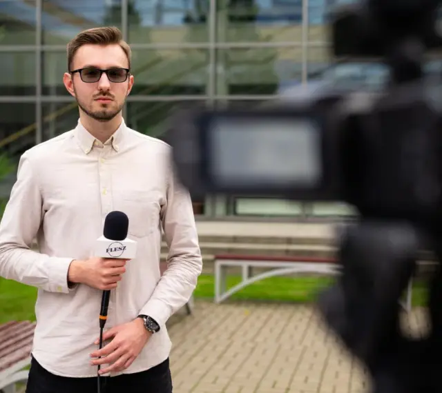 Młody dziennikarz w okularach trzyma mikrofon z logo "FLESZ" przed kamerą.