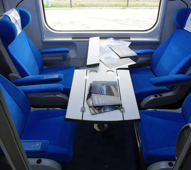 Wnętrze wagonu przedziałowego z wygodnymi, niebieskimi fotelami i stolikiem.