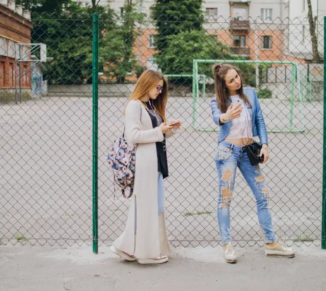 Dwie dziewczyny w modnych stylizacjach, idealnych na outfit do szkoły, stoją przy płocie. Jedna robi sobie selfie, druga przegląda telefon.