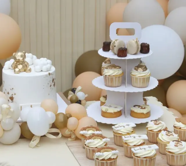 Biały tort z złotym misiem, babeczki i makaroniki na paterze, otoczone balonami w pastelowych kolorach.