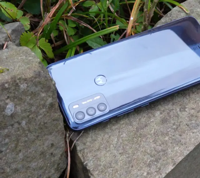 Błyszczący tył telefonu **Moto G 50** z potrójnym aparatem, leżący na kamieniach wśród zieleni.