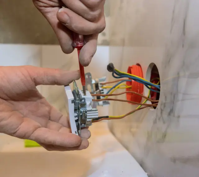 Elektryk montuje białe gniazdko, przykręcając je śrubokrętem. Widać kolorowe przewody wychodzące z puszki instalacyjnej.