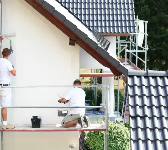 Dwóch mężczyzn na rusztowaniu maluje białą elewację domu. Sprawdź, jaka jest malowanie elewacji cena.