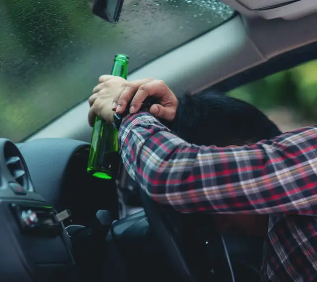 Mężczyzna w kratę trzyma butelkę, opierając głowę na kierownicy. To obraz ostrzegawczy przed jazdą pod wpływem alkoholu, przypominający o zagrożeniach i potrzebie blokady alkoholowej.