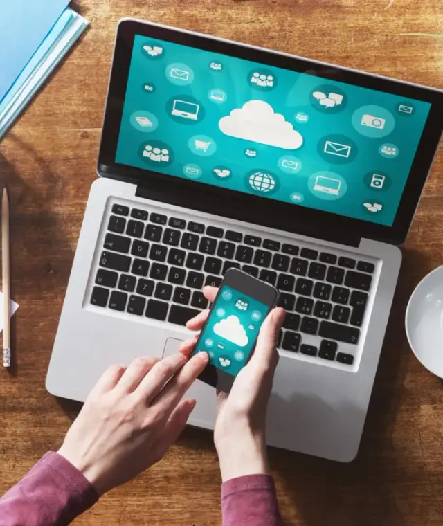 Dłonie trzymające smartfon z ikonami chmury na tle laptopa i dokumentów.