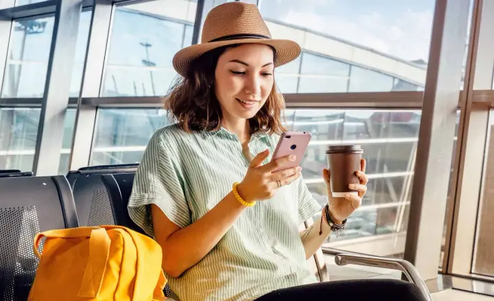 Kobieta w kapeluszu siedzi w poczekalni lotniskowej, trzymając telefon i kawę.