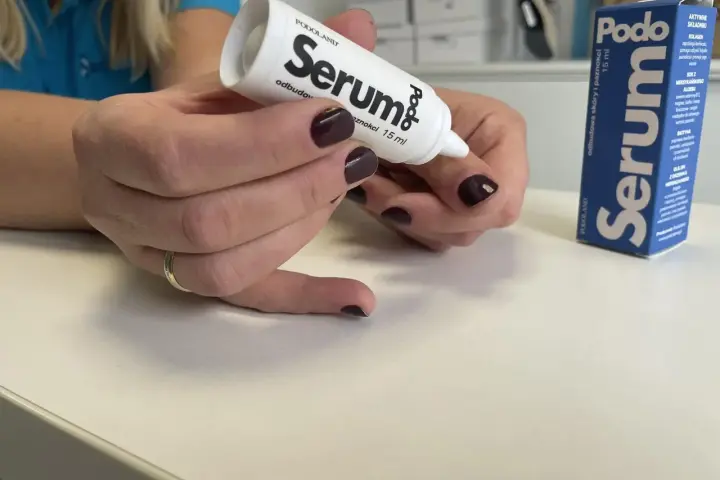 Dłonie z ciemnym lakierem aplikują podoserum na paznokieć. Opakowanie Podo Serum widoczne obok.