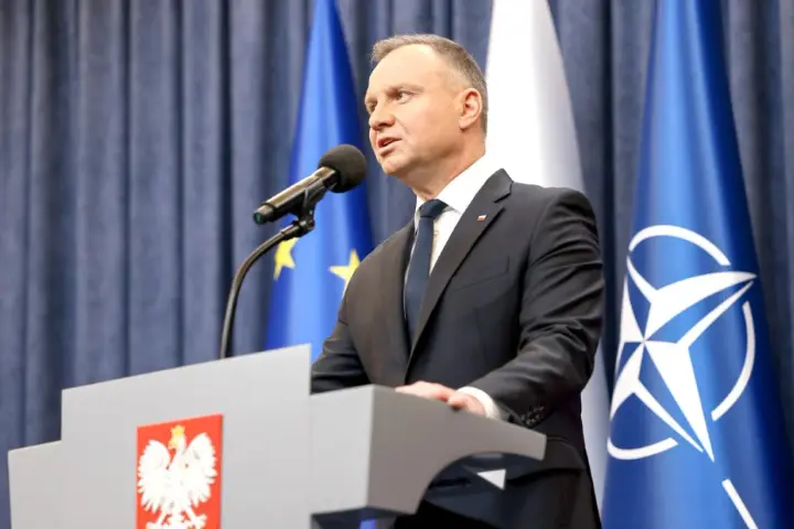 Prezydent Andrzej Duda przemawia przy mównicy z godłem Polski. Za nim flagi UE i NATO. Może to być związane z ułaskawieniem prezydenta.