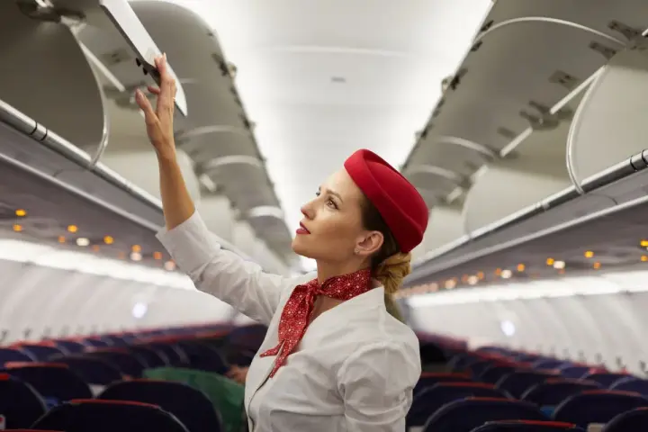 Flugbegleiterin schließt Gepäckfach. Ein Beruf, der mehr als nur das Flugbegleiter Gehalt bietet.