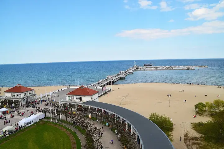 Sopot beach z molo, kawiarniami i spacerującymi ludźmi. W oddali widać statki.