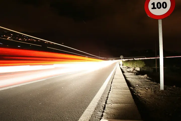 Nocna droga z długimi smugami świateł samochodowych. Znak ograniczenia prędkości 100 km/h przypomina o przepisach, jak ograniczenia prędkości w Holandii.