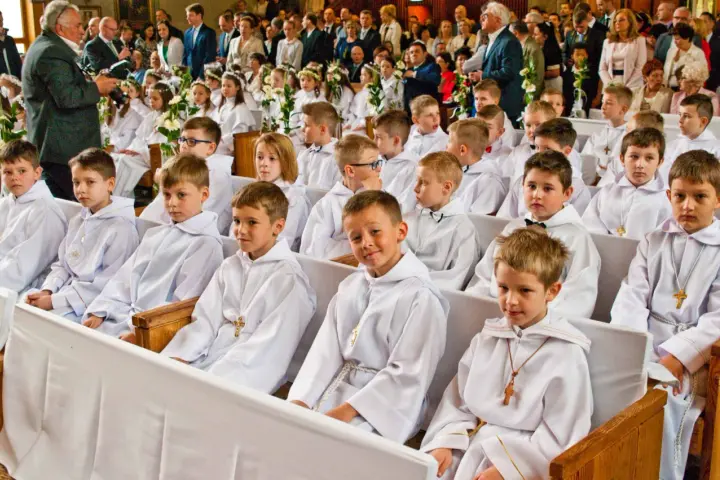 Dzieci w bieli, gotowe na rocznicę komunii świętej, siedzą w kościele. W tle widać zgromadzonych gości i fotografa.