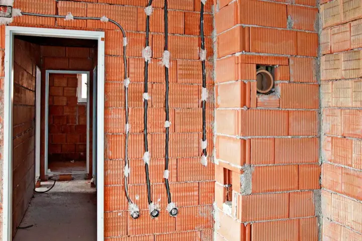 Instalacja elektryczna w trakcie budowy: łączenie kabli w ścianie bez puszki, widoczne przewody i otwory w cegłach.