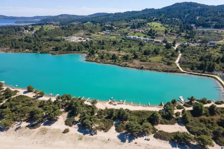 Turkusowa laguna na Kassandrze, otoczona zielonymi wzgórzami i lasem sosnowym. W oddali widać zabudowania i plażę z łodziami.