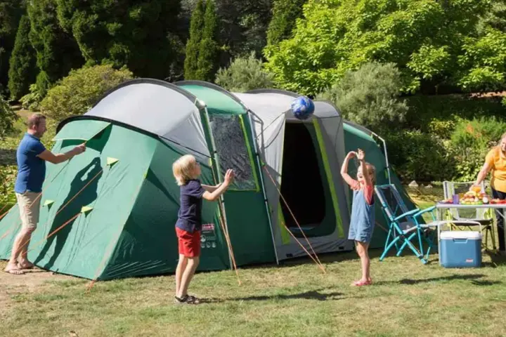 Radosny camping rodzinny: tata rozkłada namiot, dzieci bawią się piłką, mama przygotowuje posiłek.