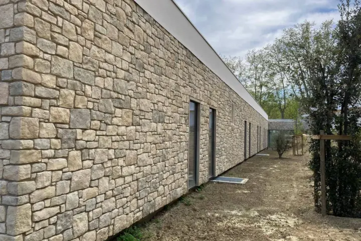 Un mur extérieur en pierres naturelles irrégulières, avec des fenêtres modernes et un toit blanc. Le sol est en terre battue.