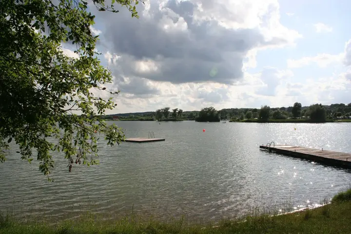 Ein sonniger Tag am Oberrieder Weiher Camping. Ein Steg mit Leiter lädt zum Baden ein, während im Hintergrund Bäume und eine idyllische Landschaft zu sehen sind.
