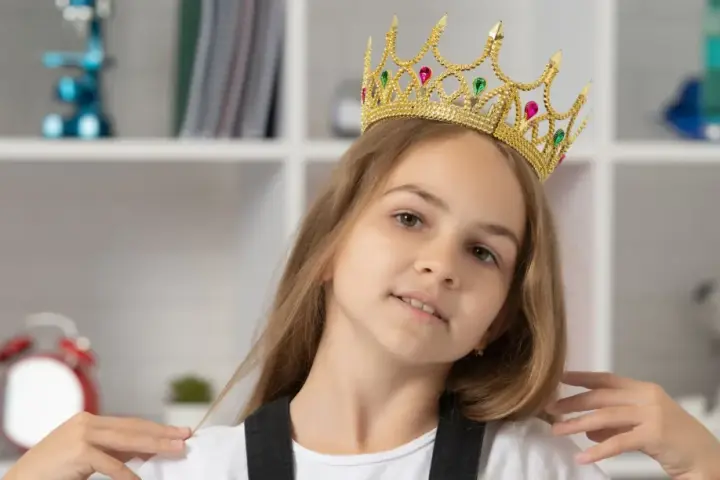 Mała egocentryczka w złotej koronie z klejnotami, pewna siebie, z uśmiechem na twarzy, na tle regału z książkami i zabawkami.