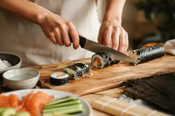 Oto jak się robi sushi: ręce kroją rolkę sushi na desce. Obok ryż, łosoś, awokado i ogórek.