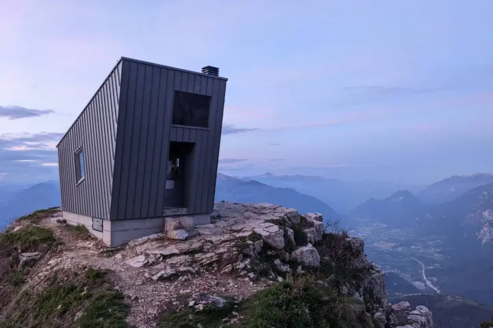 Il bivacco Giacomelli alla Madonnina, una struttura moderna su una cresta rocciosa, domina un paesaggio montano al tramonto.