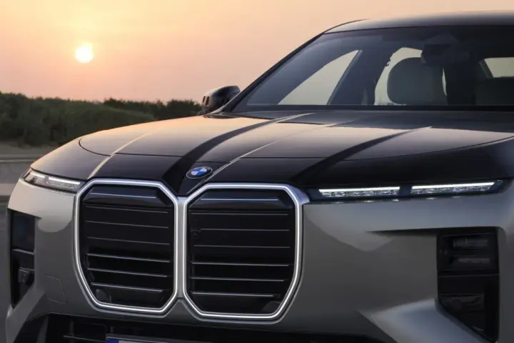 Przód luksusowego BMW M7 z podświetlaną atrapą chłodnicy i nowoczesnymi światłami, na tle zachodzącego słońca.