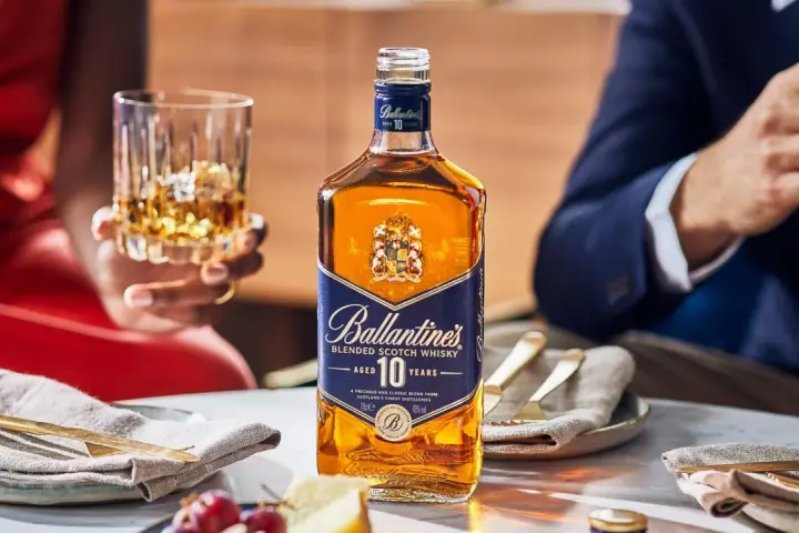 Elegancka kolacja z whisky i piwem w tle. Butelka Ballantine's 10 Years Old na stole, obok kieliszek z bursztynowym trunkiem.