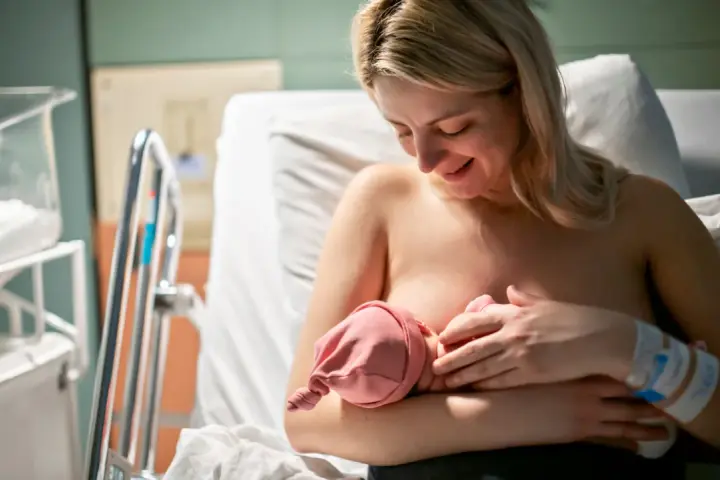 Uśmiechnięta mama karmi piersią noworodka. Pierwsze krople siary z piersi to cenny początek dla maluszka.